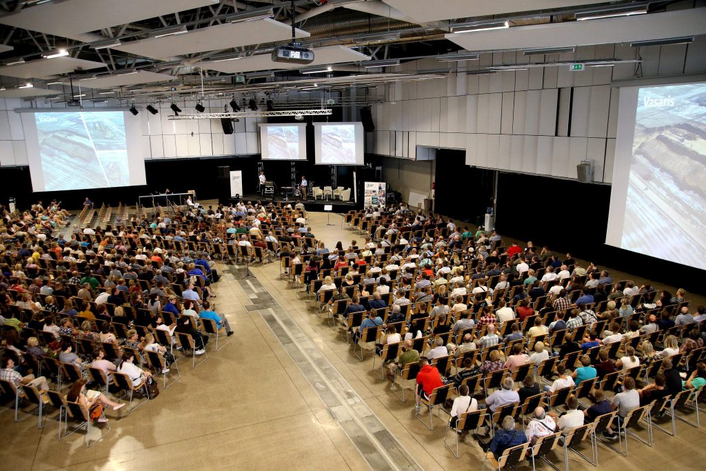 Daug žmonių sėdi ant kėdžių vienoje salėje ir stebi konferenciją